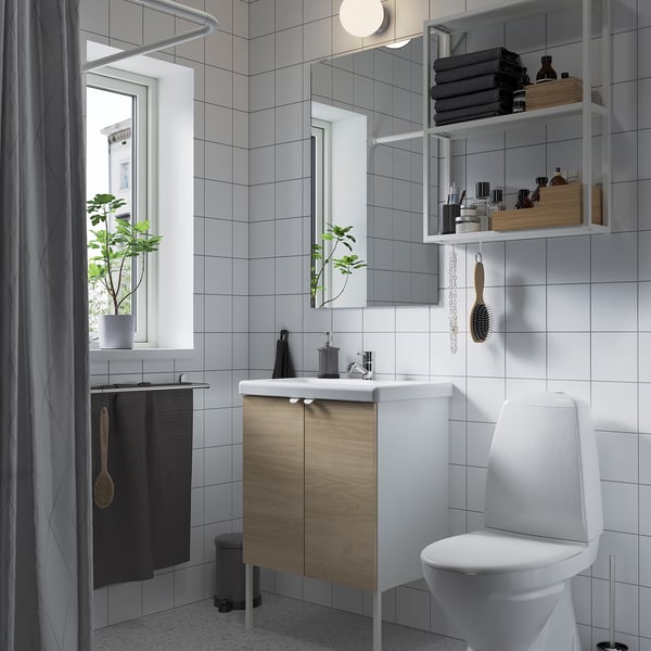 Small, modern bathroom with white tiles and a wooden vanity. A window, mirror, toilet, and open shelves.