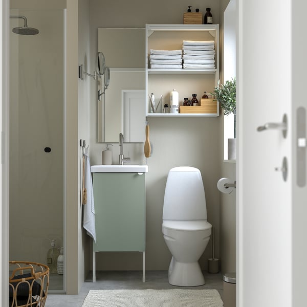 Minimalist bathroom with sage vanity, toilet, open shelves, glass shower, mirror, plants, towels.