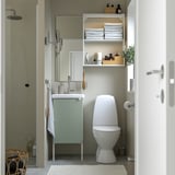 Minimalist bathroom with sage vanity, toilet, open shelves, glass shower, mirror, plants, towels.