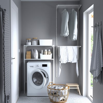 Modern grey laundry room with ENHET white washing machine, hanging shirts, shelves with laundry supplies, wicker laundry basket, and grey walls.
