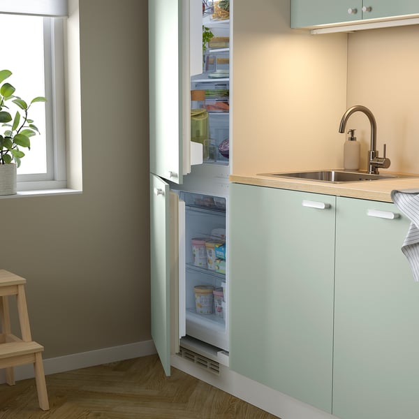 Open fridge integrated in pale green cabinet, next to sink, displaying various items.