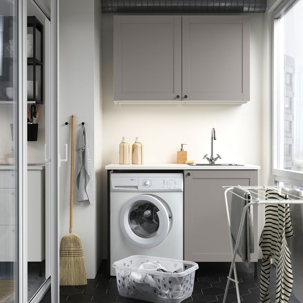 Modern compact laundry setup. Includes silver washing machine, grey cabinets, countertop sink, and hanging rack. Efficient storage solutions.