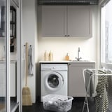 Modern compact laundry setup. Includes silver washing machine, grey cabinets, countertop sink, and hanging rack. Efficient storage solutions.