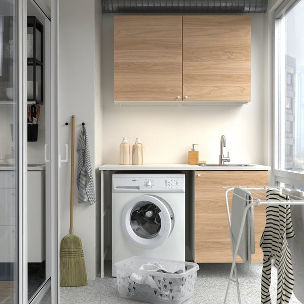 A compact laundry setup with ENHET cabinets in wood-style, white appliances, and a striped towel.