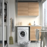 A compact laundry setup with ENHET cabinets in wood-style, white appliances, and a striped towel.