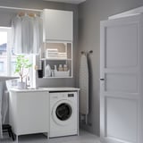 Modern laundry area with ENHET white sink, washer, & open shelves. Clothes hang nearby, with a plant & door.