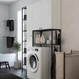 Modern laundry area with ENHET white units. Front-loading washer under black-framed shelves, towel rack to left, brown plant, black tools.