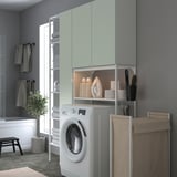 Modern grey-green laundry room with wall-mounted cabinets, white washer, open shelves, folded towels, iron, and plant.