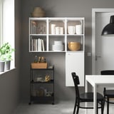Modern white ENHET kitchen unit with wall shelves, black chair, table, and black cart holding jars and baskets by window.