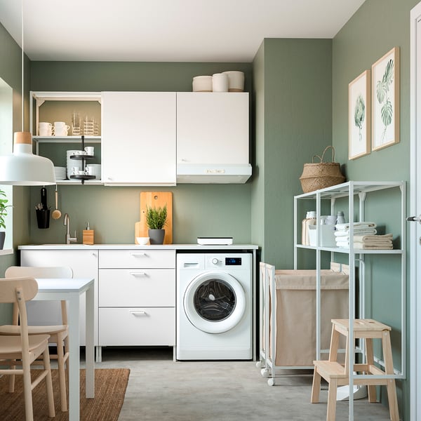 Modern kitchen with white cabinet, washing machine, dried plant, and wooden stools against green walls.