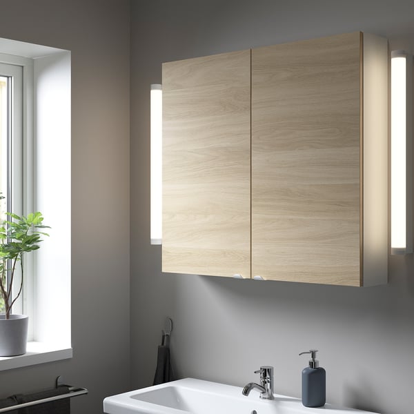 Modern bathroom with large light wood cabinet above sink, grey wall, plants by window.