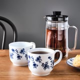 Two floral patterned mugs near a french press on a wooden table.