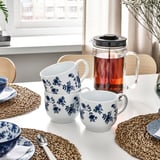 A table set with white mugs and bowls featuring blue floral patterns, a glass teapot, and woven place mats by a window.