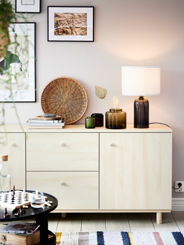 A modern light wood sideboard with clean lines, featuring drawers and cabinets, topped with a round woven tray, decorative vases, and a lamp.