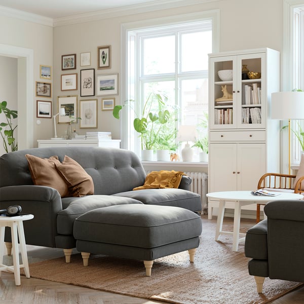 Cozy living room with ESSEBODA grey sofa, wooden feet, soft cushions, white coffee table, rug, and bookshelf with plants.