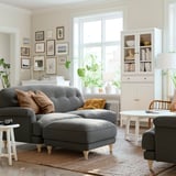Cozy living room with ESSEBODA grey sofa, wooden feet, soft cushions, white coffee table, rug, and bookshelf with plants.