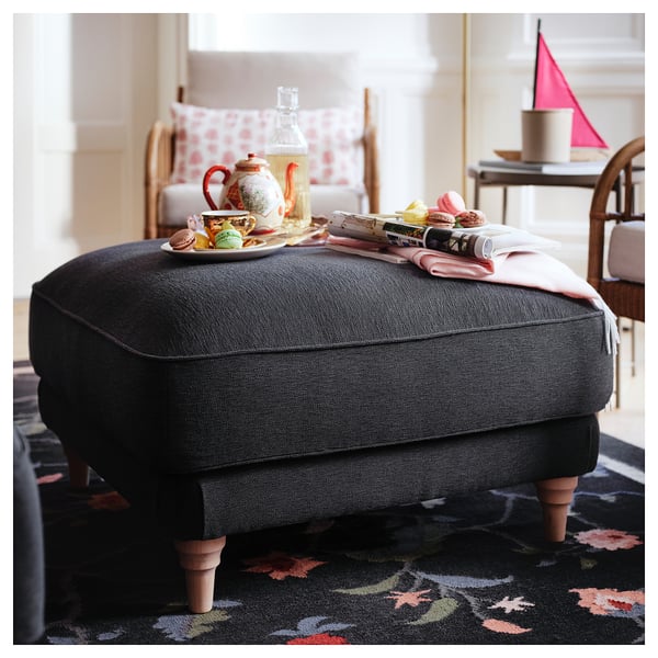 Cozy gray fabric ottoman, wooden legs, tray with tea set atop, spray of pastries, plush chair and rug.
