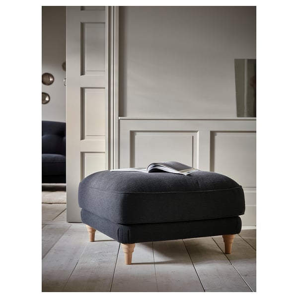 Grey fabric footstool with wooden legs on a wooden floor, against a wall.