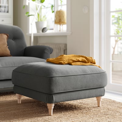 Gray footstool with wood legs, near blue sofa. Yellow blanket on top. Elegant, turned legs. Natural light streams in.