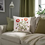 Decorative pillow named FACELIOR colorful florals on white cotton. Placed on a couch with curtains, its smaller than the cushions around it.