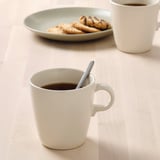 Two white mugs filled with coffee, one with a spoon, and a plate of cookies.