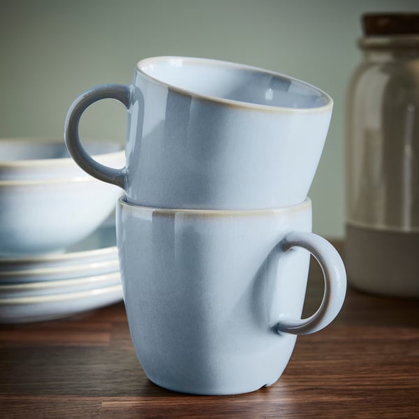 Two light blue mugs stacked on a wooden table, showcasing their simple, functional design and unique glazed finish.