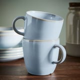 Two light blue mugs stacked on a wooden table, showcasing their simple, functional design and unique glazed finish.