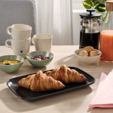 Breakfast spread with croissants on black plate, mugs, bowls, coffee press, and juice on light wood table.