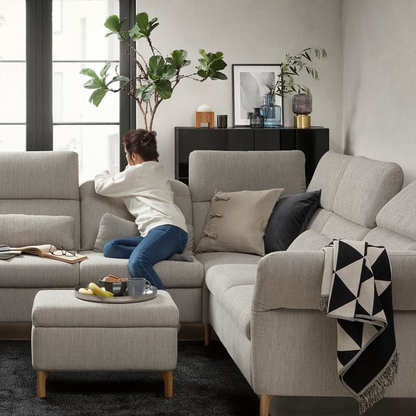 Living room scene: woman sits on grey FAMMARP sectional sofa, cushions, ottoman, black and white blanket, plants, decor, natural light.