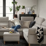 Living room scene: woman sits on grey FAMMARP sectional sofa, cushions, ottoman, black and white blanket, plants, decor, natural light.