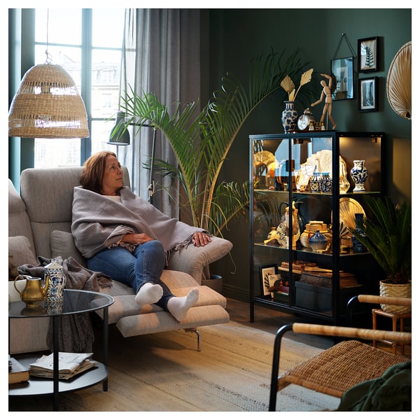 Woman relaxes on adjustable FAMMARP sofa by window, beside decorated glass cabinet and plants.