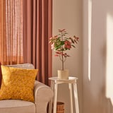 Cozy chair with mustard pillow, side table, FEJKA plant, red curtains.