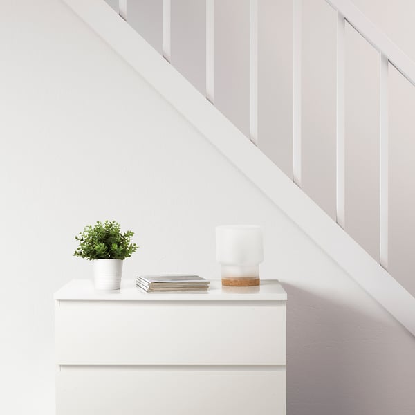 Minimalist white dresser against a staircase. Topped with a FEJKA artificial plant, stacked books, and a table lamp.
