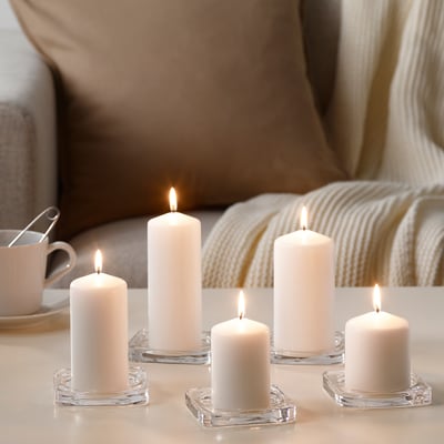 Six white candles in clear holders emit a warm glow on a table. A cozy atmosphere with a lit cup and blanket in the background.