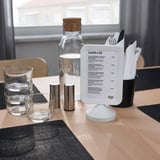 A café table set with a menu board displaying text in a frame, glasses, cutlery, napkins, and a water carafe.