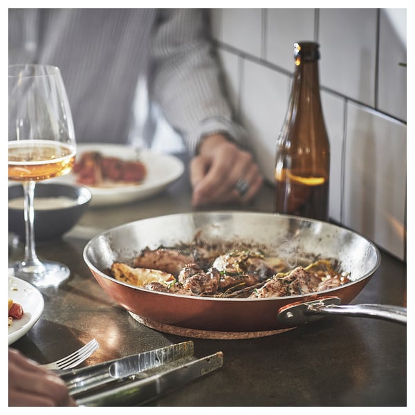 Cooking scene: copper pan with food, wine glass, beer bottle, person eating.