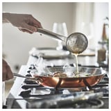 A hand pours liquid from a ladle into a copper-colored frying pan on a gas stove. Steam rises from the pan.