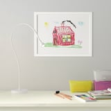 Framed childs drawing on wall, illuminated by white desk lamp. Art supplies on desk below.