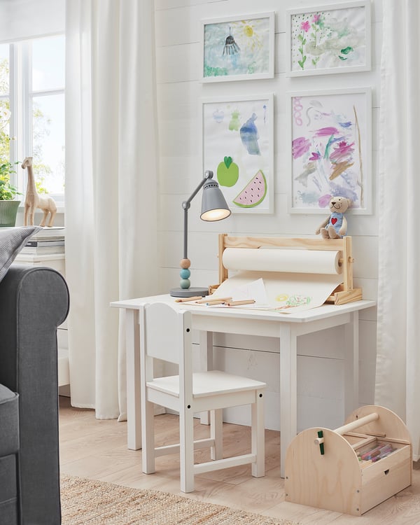 Bright kids art studio: white desk, chair, lamp, teddy bear, colorful art frames on wall.
