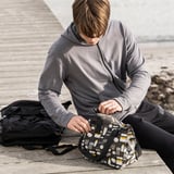 A person sits on a wooden pier unpacking a patterned black FLADDRIG lunch bag.