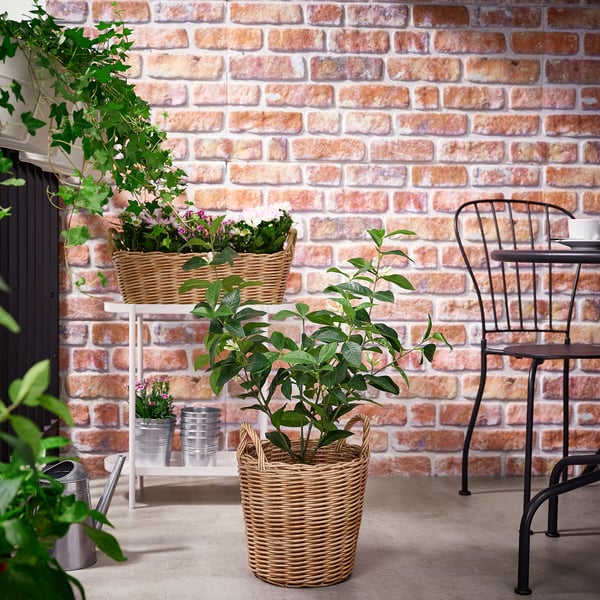 Woven FLODBJÖRK planter with green plant, black chair, white stand, brick wall.