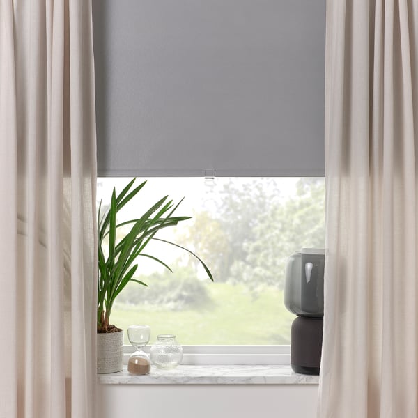Window with grey roller blind, sheer curtains, plant, vase, and speaker on sill, providing light block and modern decor.
