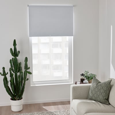 Modern living room with FÖNSTERBLAD roller blinds in light gray, fitting large window, next to large cactus, light-colored couch with green pillow.