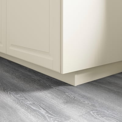White kitchen cabinet base with plinth attached, resting on gray, wood-patterned floor.