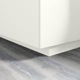 White kitchen cabinet plinth on gray wooden floor. Covers gap between floor and base cabinets. Part of FÖRBÄTTRA series.