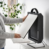 Person inserting a filter into a compact black air purifier, which improves indoor air quality and can be wall-mounted or used with a stand.