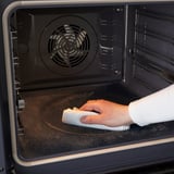 Cleaning oven interior using self-cleaning function. Hand holding cloth, wiping base.