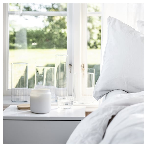 Bedside table with white candle, two glass vases, and a white pillow. A window with green outdoor view.