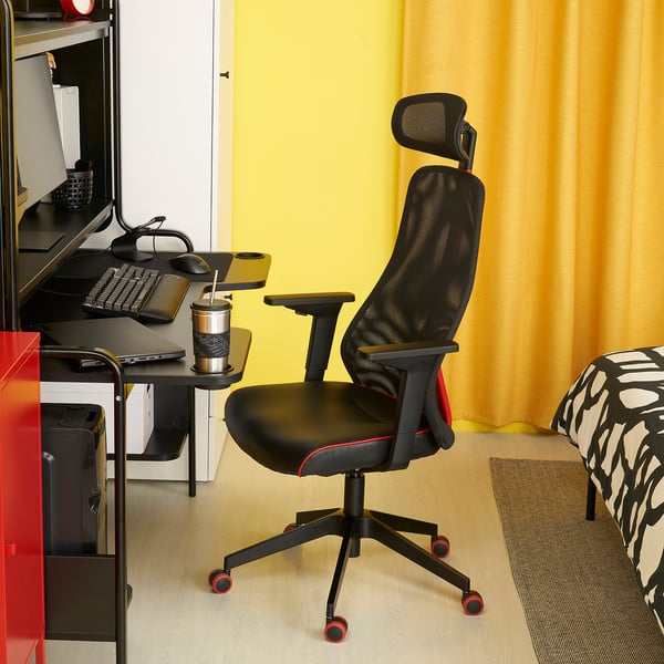 Black & red gaming chair near desk with gear, yellow curtains, patterned rug, study/nook.