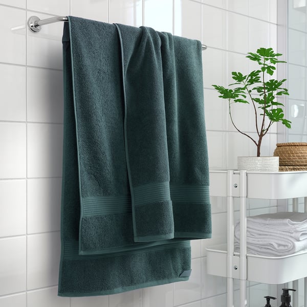 Green FREDRIKSJÖN towels on white rod, tiled wall, storage rack with extra towels and plant.